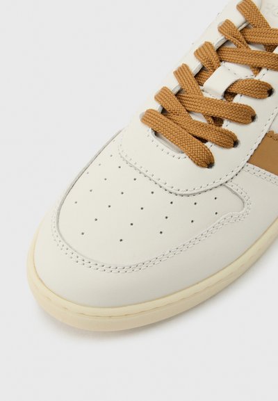 Κοντινό πλάνο ενός λευκού αθλητικού παπουτσιού με μπεζ σόλα και καφέ κορδόνια, με διατρήσεις και λεπτομερή ραφές στην περιοχή των δακτύλων.