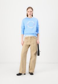 Lys blå sweatshirt med hvit tekst og sirkulært logo, sammen med beige bredbeinbukser. Svart veske og sko fullfører antrekket.