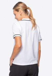Camisa polo blanca con ribetes negros en el collar y las mangas. Textura suave, mangas cortas y un ajuste relajado. Parece estar hecha de algodón.