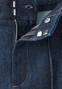 Gros plan sur un jean en denim bleu foncé montrant la braguette ouverte, la fermeture éclair métallique, le bouton et les agrafes avec coutures visibles.