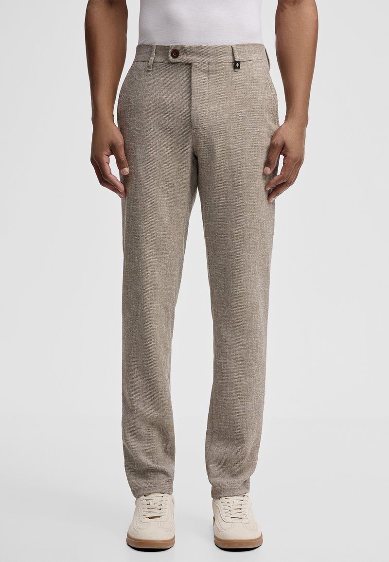 Man draagt beige textuurbroeken, witte sneakers en een wit overhemd, staand tegen een effen lichte achtergrond.