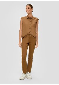 Chemise sans manches brun avec poche avant et pantalon slim fit brun assorti, en tissu texturé, accompagnés de baskets blanches.