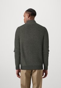 Dark green ribbed sweater with a high collar and long sleeves, featuring a textured knit design. Paired with light-colored trousers.