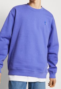 Felpa viola con colletto rotondo e polsini a coste. Presenta un piccolo logo di un uccellino blu sul petto. Indossata sopra una camicia bianca a strati.
