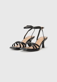 Schwarze High Heels Sandalen mit mehreren dünnen Riemen, offener Zehenpartie und einem moderaten Blockabsatz. Glattes, lederähnliches Material mit einer beigen Innensohle.