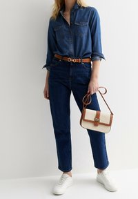 Kvinna iklädd en mörkblå denimskjorta och jeans med brunt bälte, håller en beige och brun handväska, och bär vita sneakers.