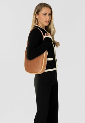 Vrouw in zwart-witte cardigan houdt een tan leren schoudertas met geperforeerd cirkelvormig logo vast, staand tegen een effen lichte achtergrond.