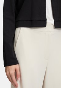 Kobieta w kremowych spodniach o wysokim stanie i czarnym długim topie na krótki rękaw, z lewą ręką swobodnie opuszczoną wzdłuż ciała.