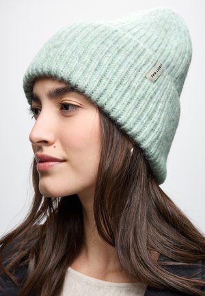 Cappello di maglia verde con una trama a costine e un leggero slouch, dotato di un piccolo etichetta sul lato. Materiale morbido e caldo.