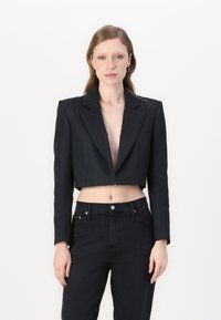 Zwarte cropped jas met lange mouwen, reverskraag, textuurstof en open voorkant. Gecombineerd met hoge zwarte jeans.