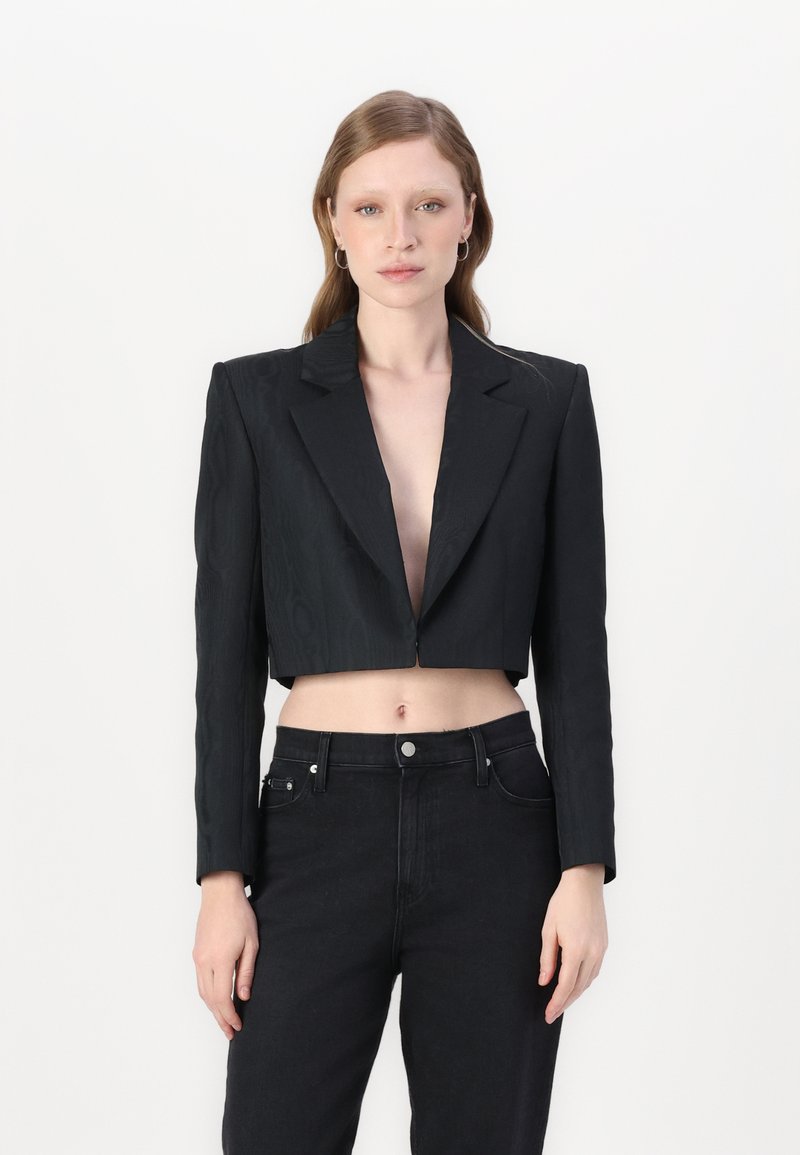 Zwarte cropped jas met lange mouwen, reverskraag, textuurstof en open voorkant. Gecombineerd met hoge zwarte jeans.