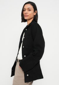 Μαύρο denim μπουφάν με κούμπωμα με κουμπιά, τσέπες στο στήθος και άνετη εφαρμογή, συνδυασμένο με λευκό τοπ και μπεζ παντελόνι. Ομαλή υφή.