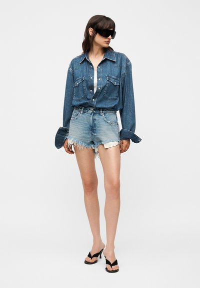 Γυναίκα που φοράει υπερμεγέθη τζιν πουκάμισο, ανοιχτόχρωμα ξεφτισμένα τζιν σορτς, μαύρα σανδάλια με ψηλό τακούνι και μεγάλα μαύρα γυαλιά ηλίου, στέκεται μπροστά σε λευκό φόντο.