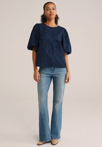 Top azul marino bordado con mangas abullonadas, que presenta un patrón texturizado y ondulado, combinado con pantalones vaqueros flare de color azul claro y zapatos beige.