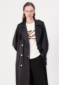 Cappotto nero con ampi risvolti, design doppiopetto e bottoni argentati; indossato sopra una camicia crema con un logo marrone.