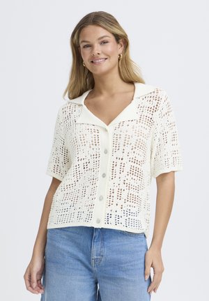 Žena sa usmieva, oblečená v bielom krátkom rukáve háčkovanom topíku s gombíkmi a golierom, v svetlomodrých džínsoch, stojaca pred jednoduchým pozadím.