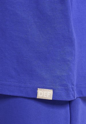 Plavi pamučni majica s glatkom teksturom, kratkim rukavima i pravokutnom oznakom od tkanine s natpisom "DEF" blizu rubova.