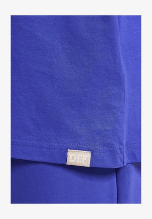 Plavi pamučni majica s glatkom teksturom, kratkim rukavima i pravokutnom oznakom od tkanine s natpisom "DEF" blizu rubova.