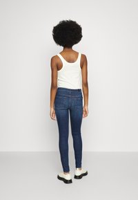 Jeans ajustados de mezclilla azul oscuro, con una cintura alta, bolsillos traseros y un acabado suave, combinados con una camiseta sin mangas blanca y zapatos negros.
