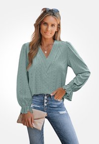 Miętowo-zielona bluzka w serek z koronkowymi detalami, bufkowymi rękawami i luźnym krojem, zestawiona z niebieskimi dżinsami z wysokim stanem i beżową torebką.