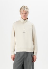 Bežna zip-up dukserica s visokim ovratnikom i elastičnim manžetama. Sadrži mali crni logo na prsima. Nosena je s sivim hlačama.