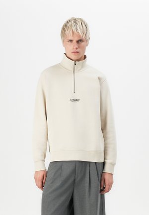 Beige zip-up sweatshirt with a high collar and elastic cuffs. Features a small logo in black at the chest. Worn with gray trousers.