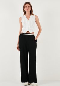 Bela srajčna brezrokavnika s vertikalnimi črnimi črtami, v kombinaciji z ohlapnimi črnimi hlačami in belimi športnimi čevlji. Preprost, krojen dizajn.