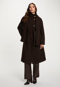 Kvinde iført en mørkebrun bæltejakke med store knapper, matchende tørklæde draperet over den ene skulder, brune bukser og ankelboots med hæl.