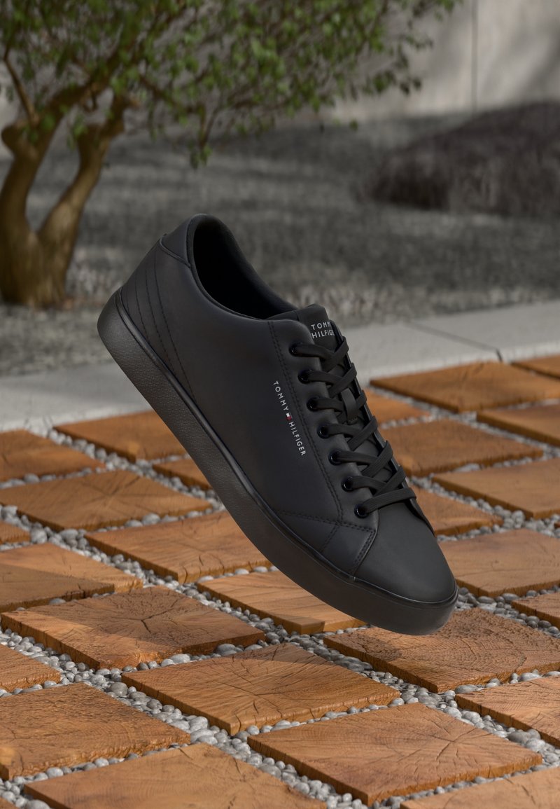 Baskets noires Tommy Hilfiger avec lacets sur des carreaux en bois carrés avec des cailloux blancs, une plante verte en arrière-plan flou.