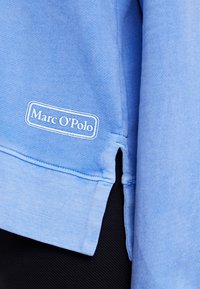 Sudadera azul de algodón con cuello redondo, dobladillo cosido, aberturas laterales y un logo blanco bordado de "Marc O'Polo" en la parte inferior izquierda.