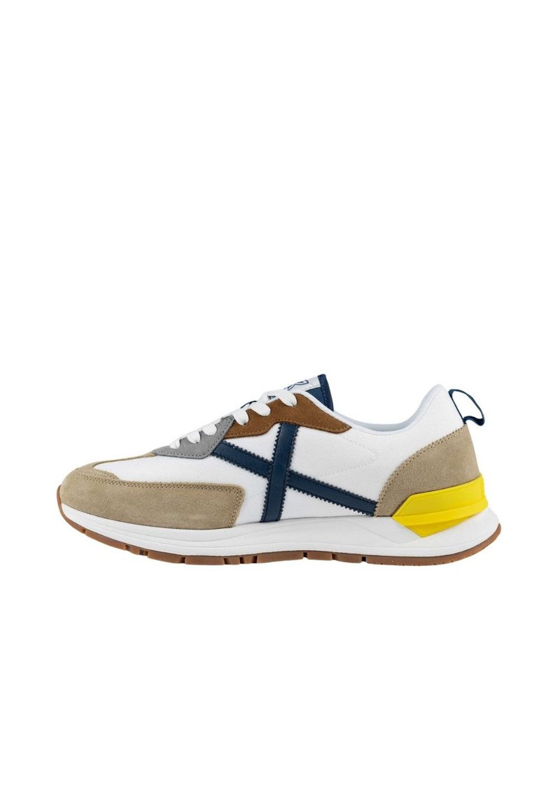 Zapatilla blanca con acentos en gris, marrón y azul marino. Hecha de ante y malla, presenta una franja lateral prominente y un detalle en el talón en color amarillo.