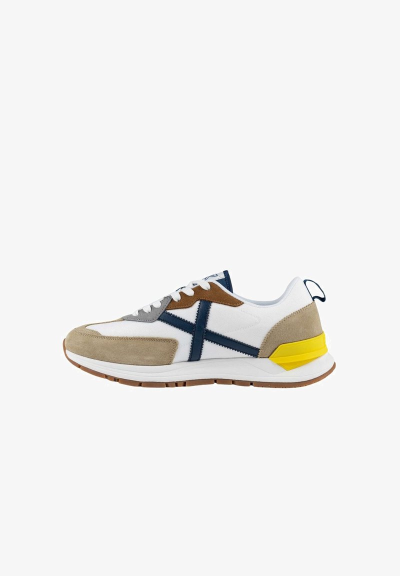 Zapatilla blanca con acentos en gris, marrón y azul marino. Hecha de ante y malla, presenta una franja lateral prominente y un detalle en el talón en color amarillo.