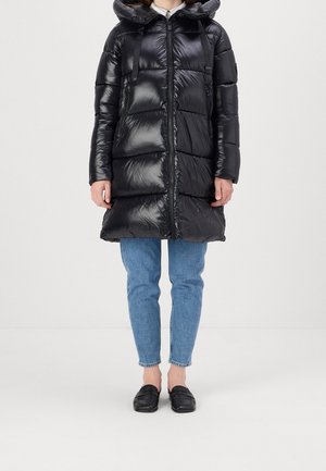 Persoon gekleed in een lange zwarte donsjas met capuchon, blauwe spijkerbroek en zwarte instapschoenen, staand tegen een effen achtergrond.