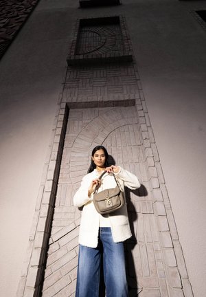 Beigelederhandtasche mit einem Verschluss, gehalten von einer Person in einem weißen, flauschigen Mantel und blauen, weiten Jeans, vor einer strukturierten Wand.