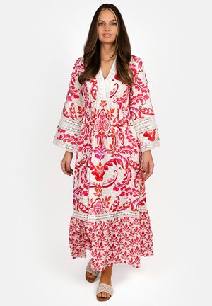 Woman in a white and pink floral patterned long dress with wide sleeves, walking forward on a plain white background.