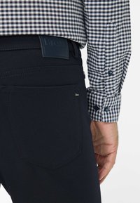 Marineblaue Hose mit einer Rückentasche und einem blauen Lederetikett. Dazu wird ein blau-weiß kariertes Hemd mit Knopfdetails getragen.