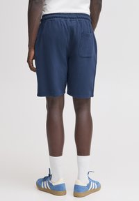 Tmavomodré bavlnené šortky s elastickým pásom a zadným vreckom, s voľným strihom a olemovanými hranami. Nosia sa s modrými teniskami a bielymi ponožkami.