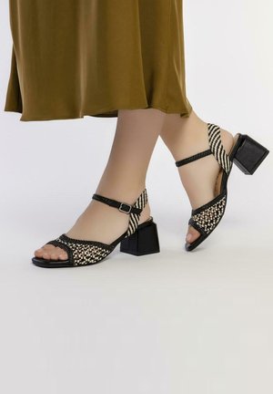 Sandales à talon tressées en noir et blanc avec un talon carré, orteils ouverts et une bride au cheville. Elles présentent un matériau texturé et un motif géométrique.