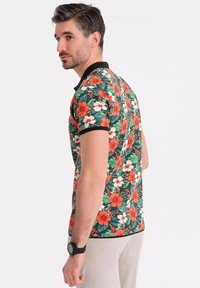 Blomstret polo-shirt i grøn med orange og hvide blomster, sort krave og en tætsiddende pasform. Model iført lyse bukser og et ur.