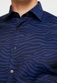 Blå skjorta med knappar i en strukturerad mörkblå tyg med ett vågigt randmönster. Fyra knappar synliga vid knappslån. Skräddarsydd krage.