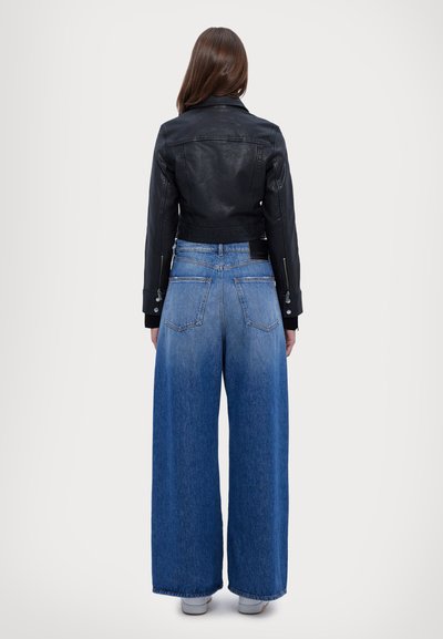 Woman standing with back facing camera wearing black cropped leather jacket, wide-leg blue jeans, and white shoes on plain background.