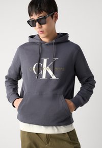 Siva kapuca s sprednjim žepom, z belim in zlatim logotipom 'Calvin Klein Jeans'. Nosena preko belega sloja z elastičnimi manšetami.