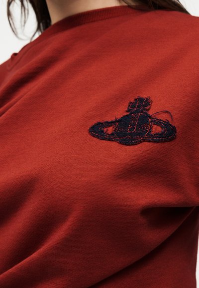 Κοντινό πλάνο ενός πουκαμίσου χρώματος σκουριάς με ένα υφασμάτινο μαύρο κεντητό έμβλημα σφαίρας και δαχτυλιδιού στο στήθος.