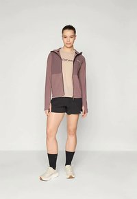 Lett purpurfarget jakke med hette, med glidelås og kontrasterende ermer. Bært over en beige skjorte og svarte shorts.