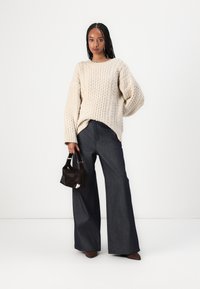 Dicker, creamfarbener Strickpullover mit überschnittenen Schultern, kombiniert mit weit geschnittenen, dunkelblauen Jeans. Hält eine kleine schwarze Handtasche.