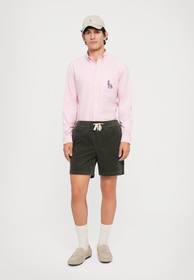 Chemise boutonnière rose clair avec un motif brodé, associée à des shorts en velours côtelé vert foncé, des chaussettes blanches et des mocassins beiges.