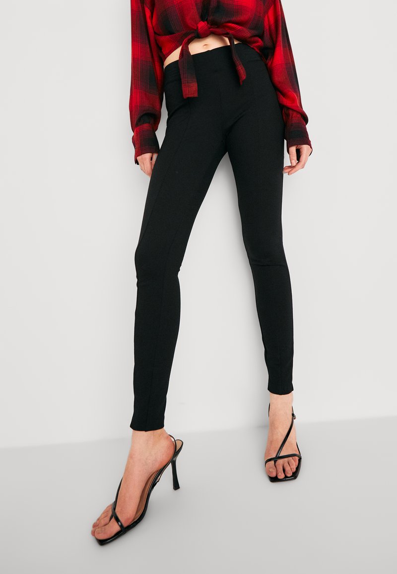 Leggings noirs avec une taille haute, un design ajusté et des détails de couture latérale, associés à une chemise à carreaux rouges et des sandales à talons noires.