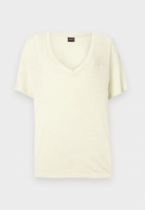 Tricou crem cu mâneci scurte și decolteu în V, cu o etichetă neagră mică cu text roșu la nivelul gâtului.