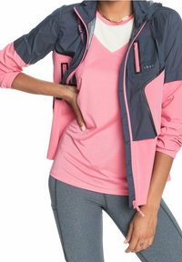 Giacca a vento rosa e blu navy dal design sportivo, con tasche con zip e un tessuto leggero, indossata sopra un top da corsa rosa.