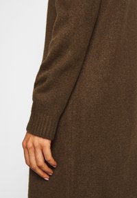 Mano appoggiata di lato, che indossa un indumento lungo a maniche con maglia a coste marrone, su uno sfondo chiaro e semplice.